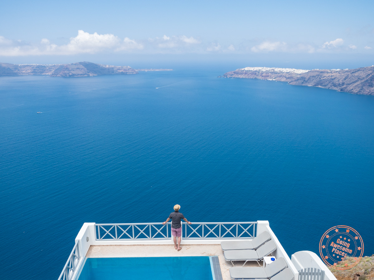 A person stands at the edge of a balcony overlooking a deep blue sea of Santorini, with an infinity pool in the foreground. The coastline and distant islands are visible under a clear sky. Two lounge chairs are on the right side of the pool.