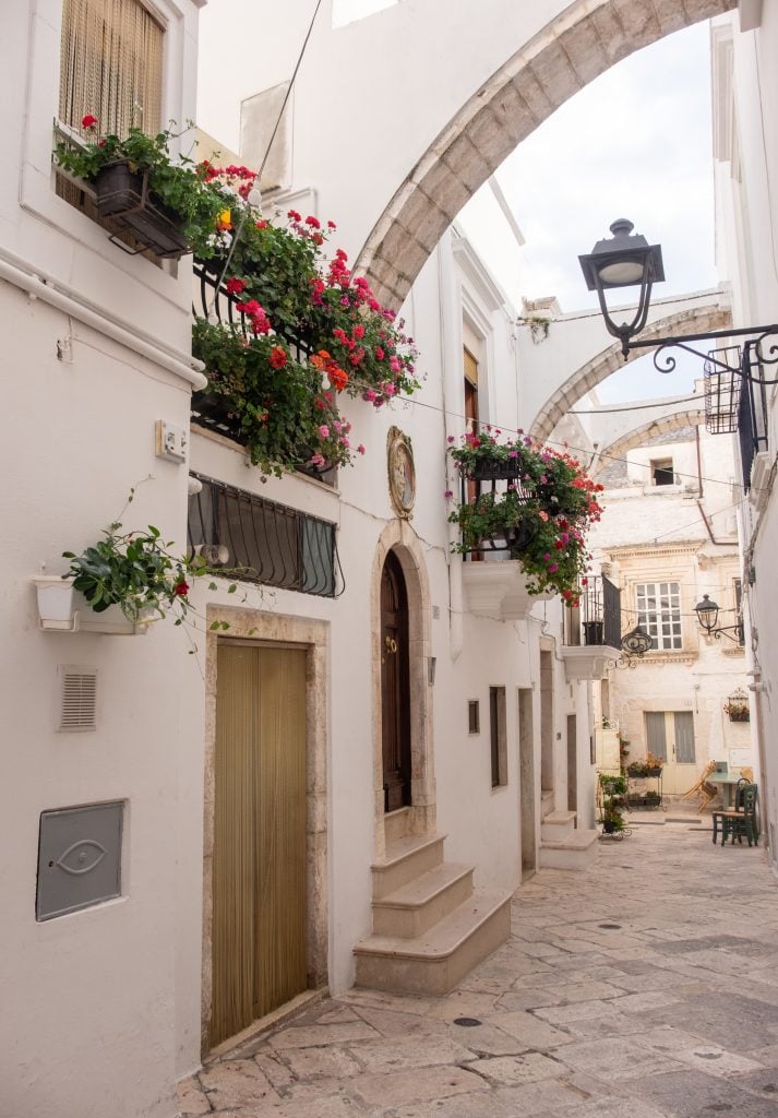 An alleyway with white walls and geraniums dangling from windows in Italy.