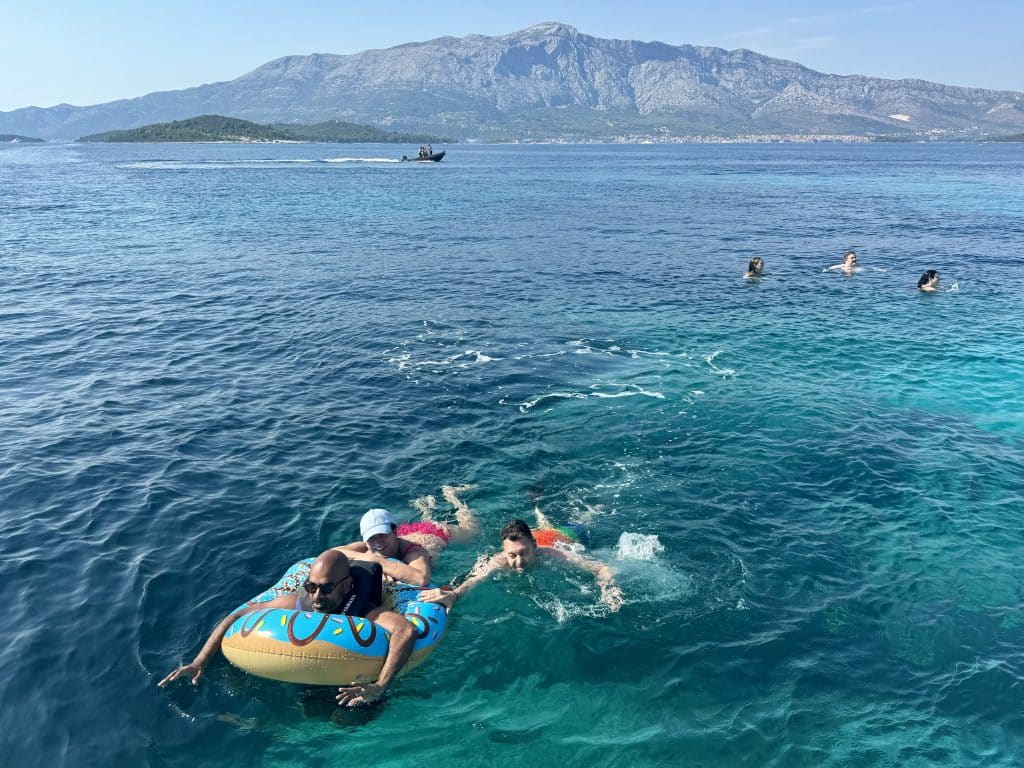 A big group of friends playing in the bright blue clear water, Kash in an inner tube, people pushing him.