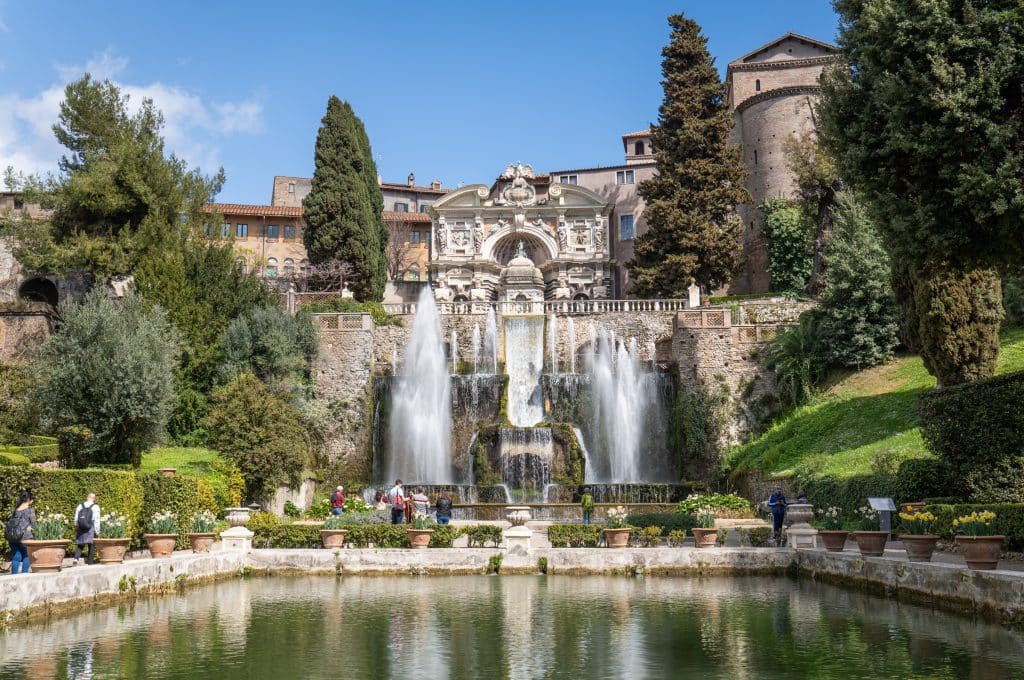 A big, ornate outdoor fountain with an emerald green pool in front of it. People are walking around and taking pictures.