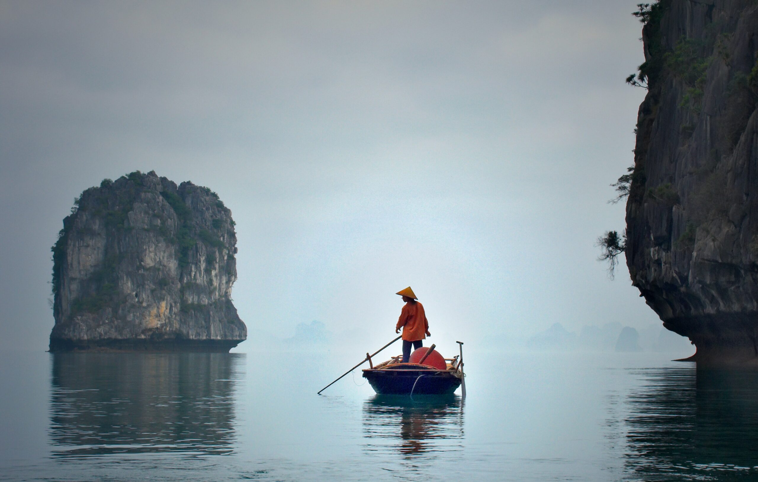 A fisher in a small boat fishes in the morning mist near two rocky islets