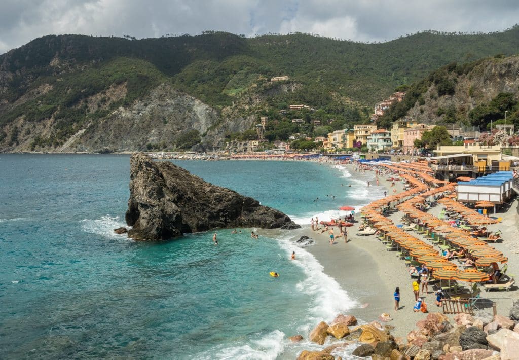 A seaside town in Italy with a Long Beach covered in orange and green-striped umbrellas. There's a big boulder hanging out in the clear teal water, and you can see green mountains rising in the background.