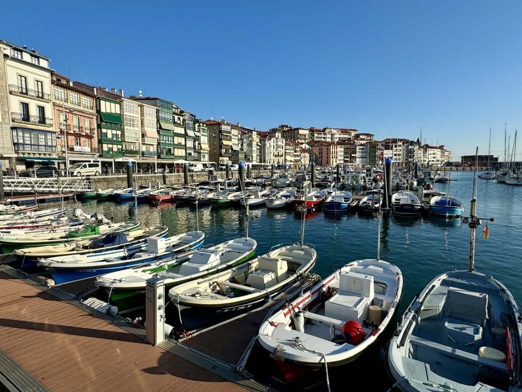 Fishing boats lined up in a calm harbor in the Basque Country, with square buildings with large windows in the background.