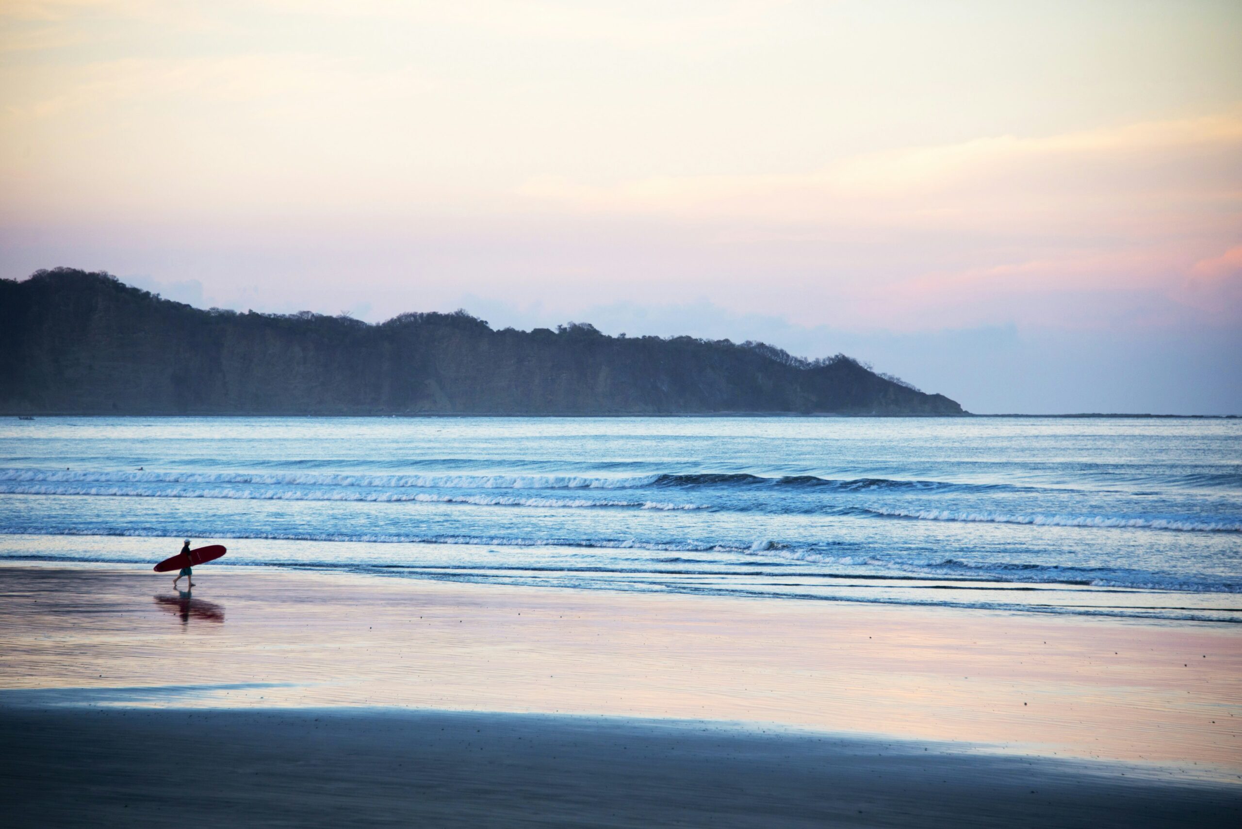 A person walks on a beach in Nosara at sunset, surfboard in hand