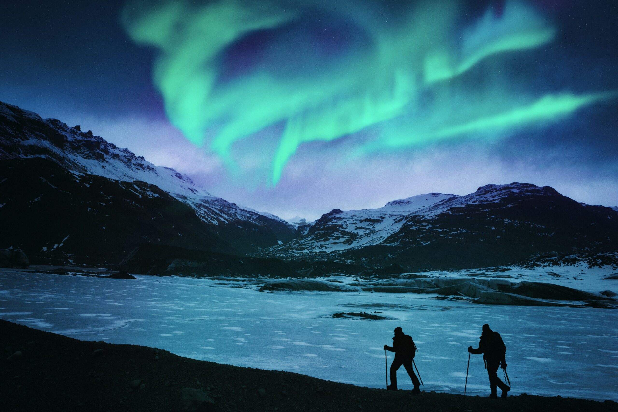 Two hikers in silhouette follow the edge of a frozen body of water at night as the northern lights streak the sky green and purple above them