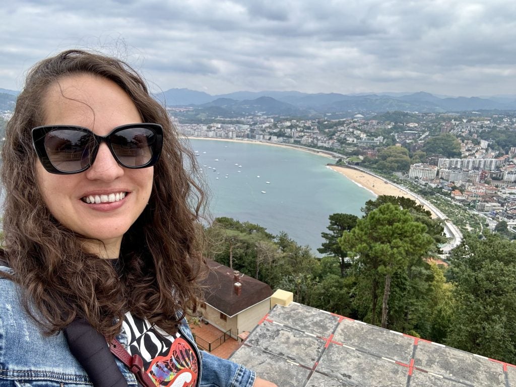 Kate taking a smiling selfie in sunglasses in front of a view of a long, curved beach, on a cloudy day.