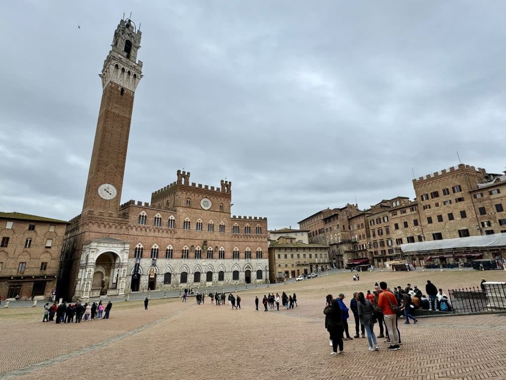 A big town square in Italy with reddish bricks on the pavement, surrounded by red brick buildings, including a tall clock tower.