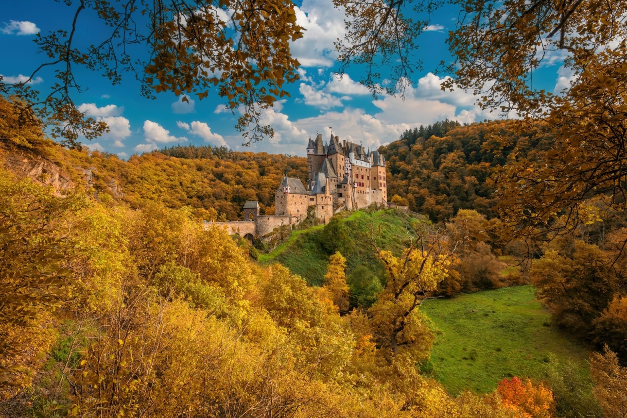 Majestic Burg Eltz castle surrounded by vibrant fall foliage in Rhineland-Palatinate, Germany. Medieval fortress perched on a hill, framed by golden trees under a blue sky.