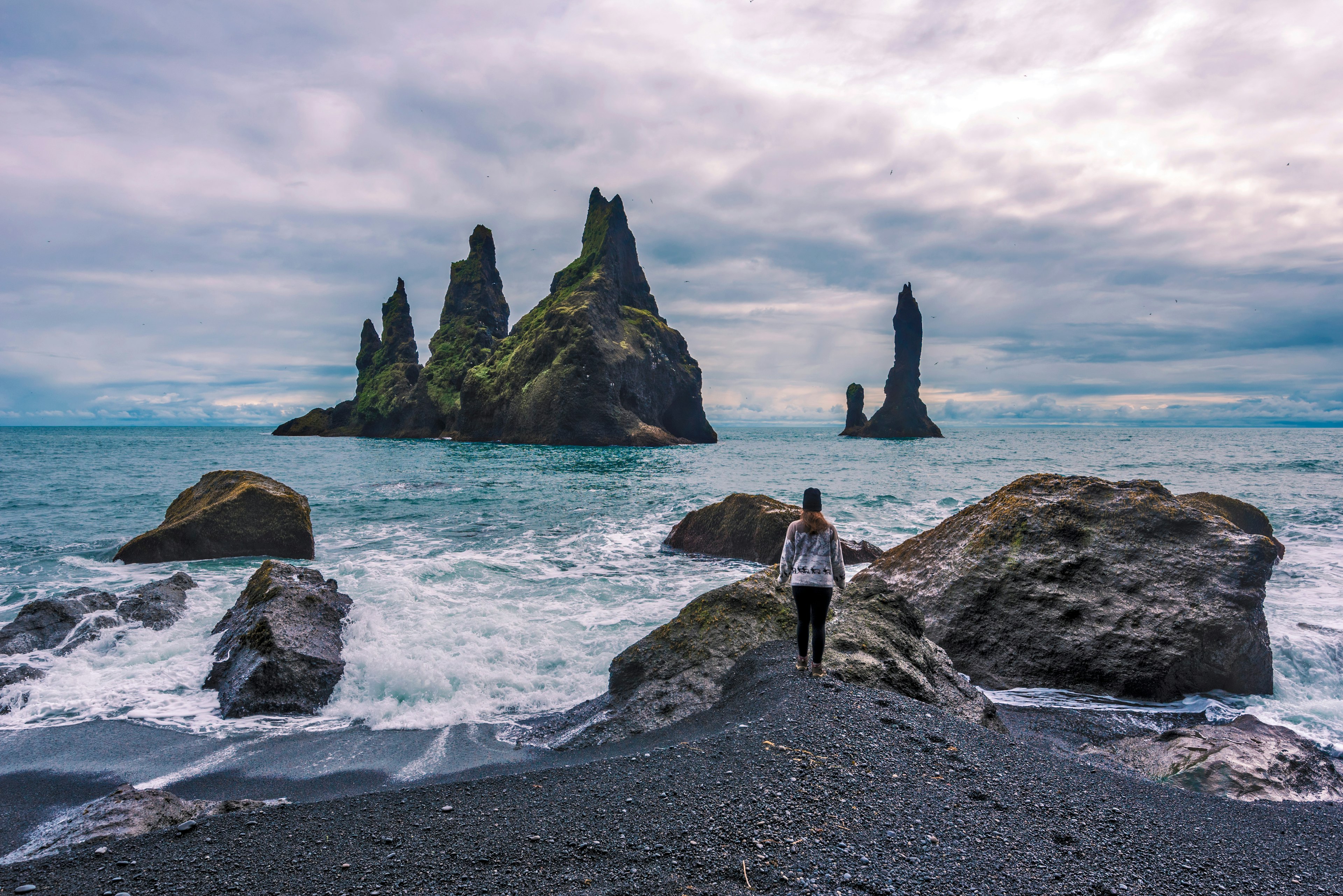 A woman stands on the rocks of a wild beach with several jagged stacks out at sea