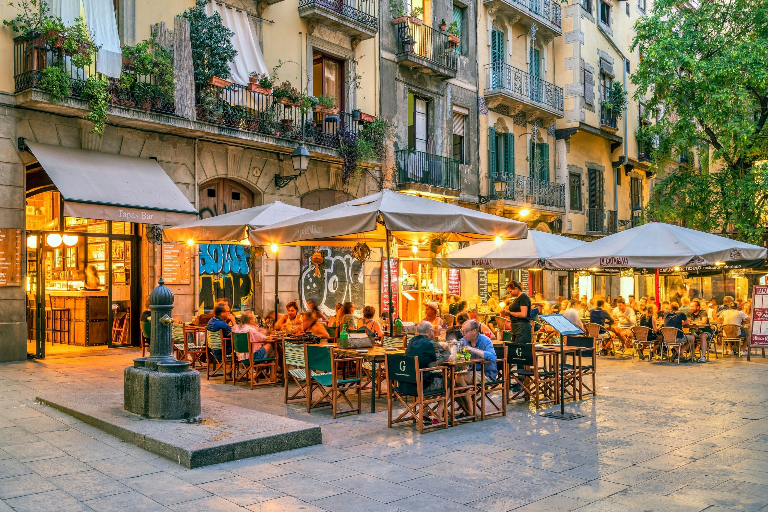 People sit at tables under umbrellas on a pedestrianized street, lit by the glow of restaurants at dusk