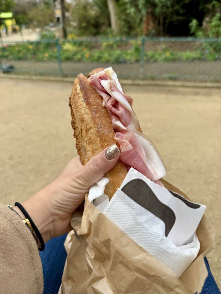A hand holding a ham sandwich on a baguette in a park