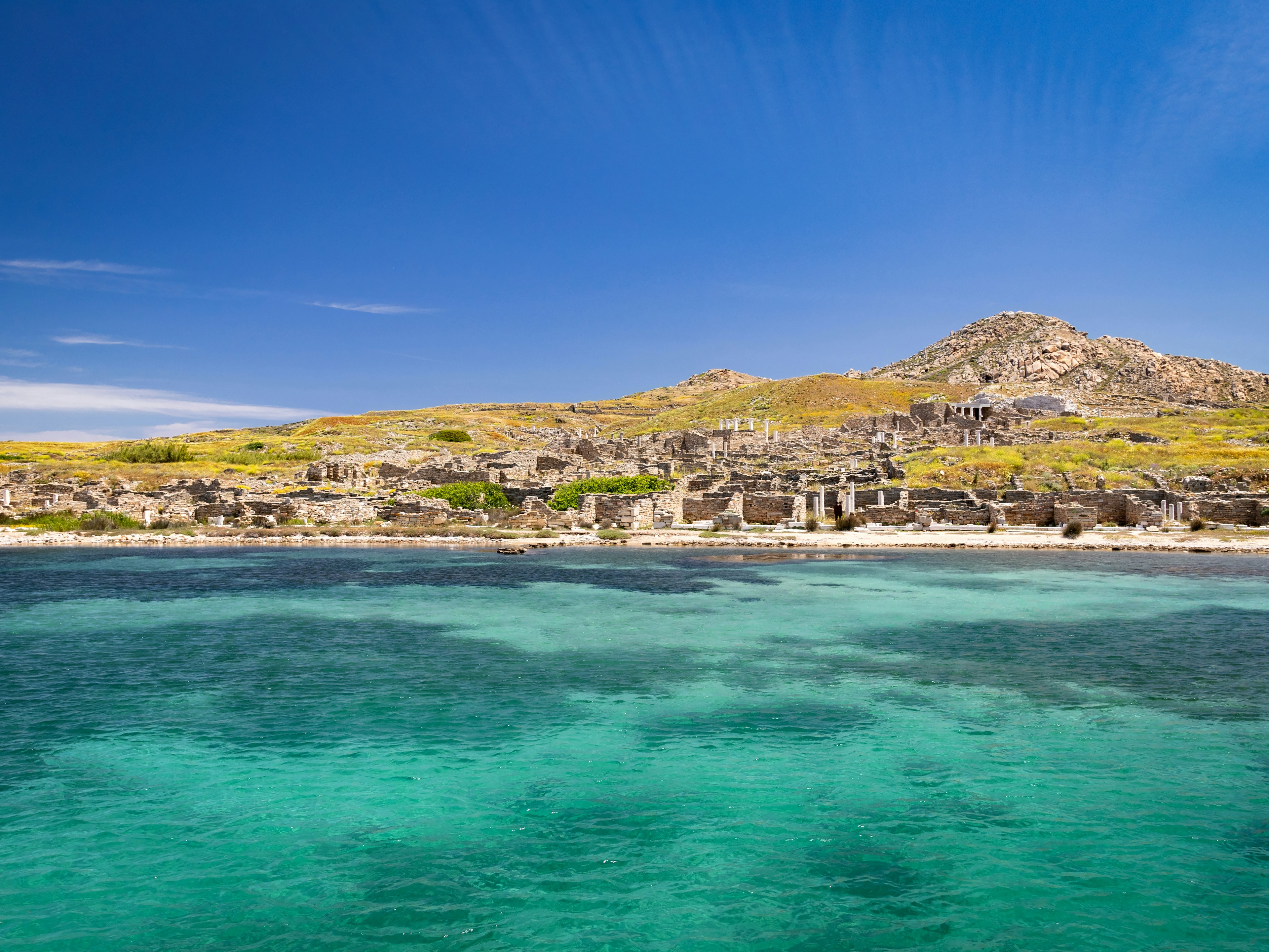 A view of the ancient ruins on the island of Delos with crystal clear blue water in the foreground.