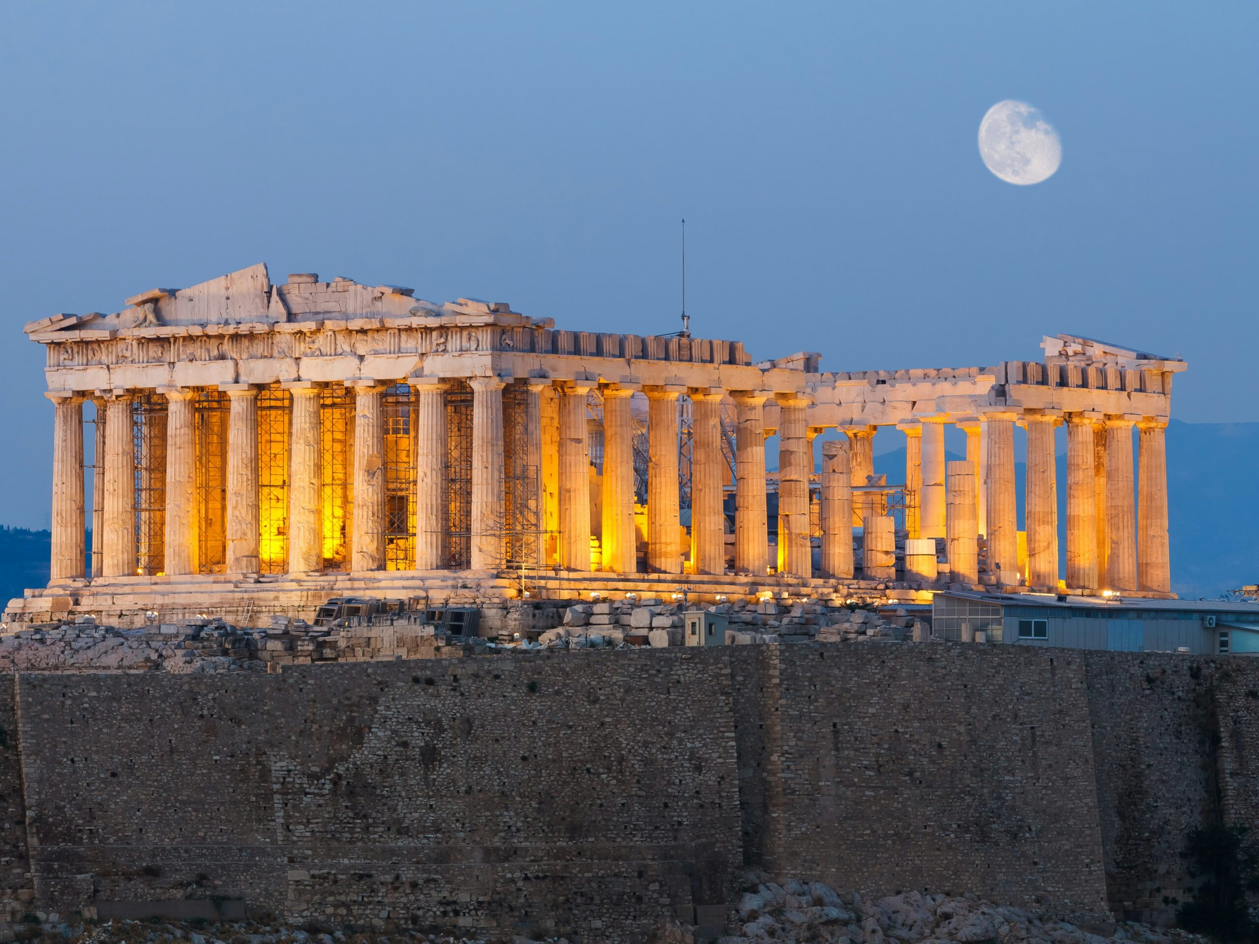 The lit-up structure of the Parthenon at night, with the moon in the background.