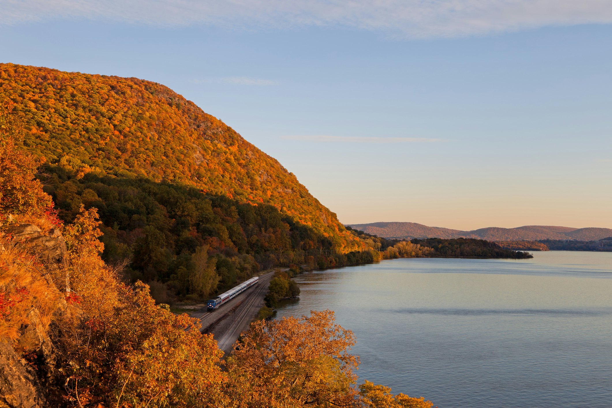 Fall foliage surrounding a NYC commuter train at sunset along the Hudson River in the Hudson Valley.