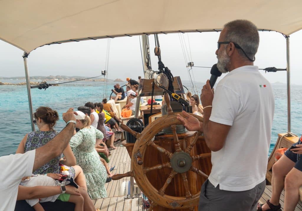 A captain behind the wheel of a ship, lots of people looking at the sea.