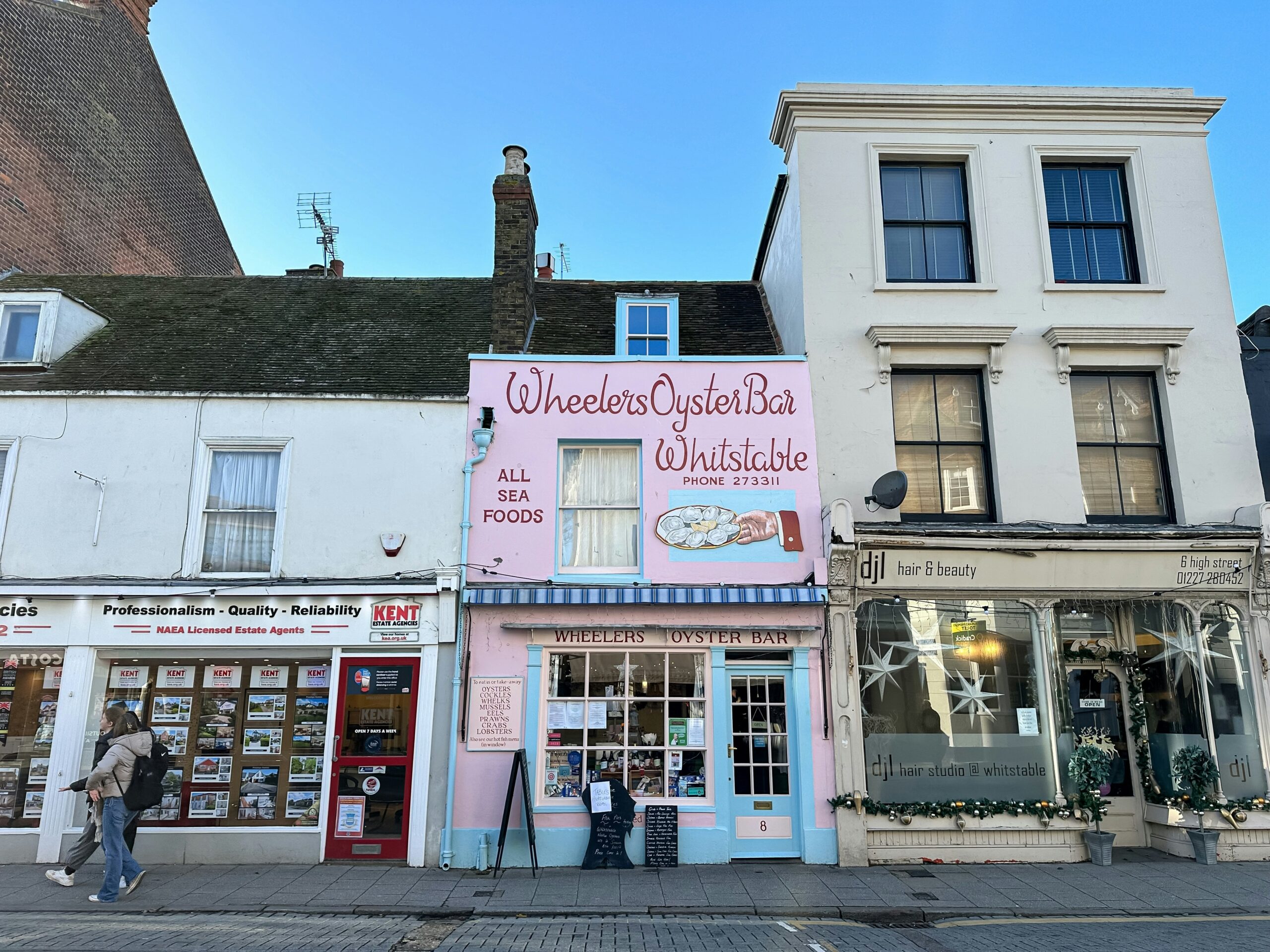 A two-story building painted pink, with the name Wheelers Oyster Bar Whitstable in red script and a light blue doorway, between two beige buildings.