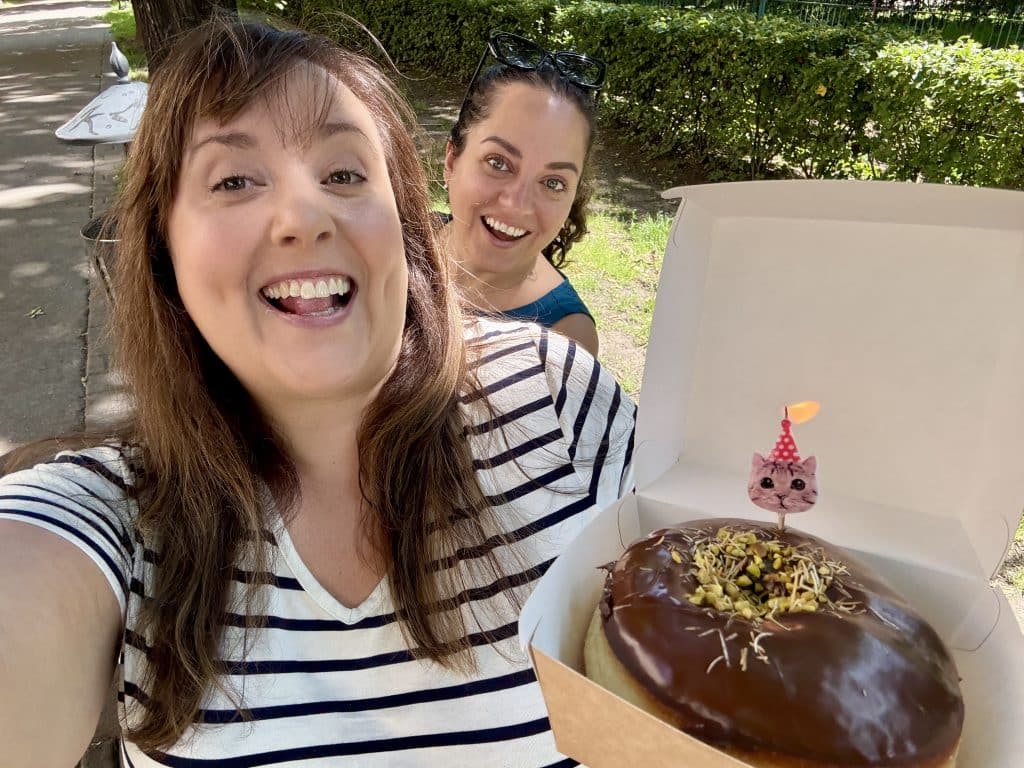 Cailin and Kate take a selfie on a park bench. Cailin holds a paczki, a Polish-style donut topped with chocolate and pistachio with a cat-shaped birthday candle on top.