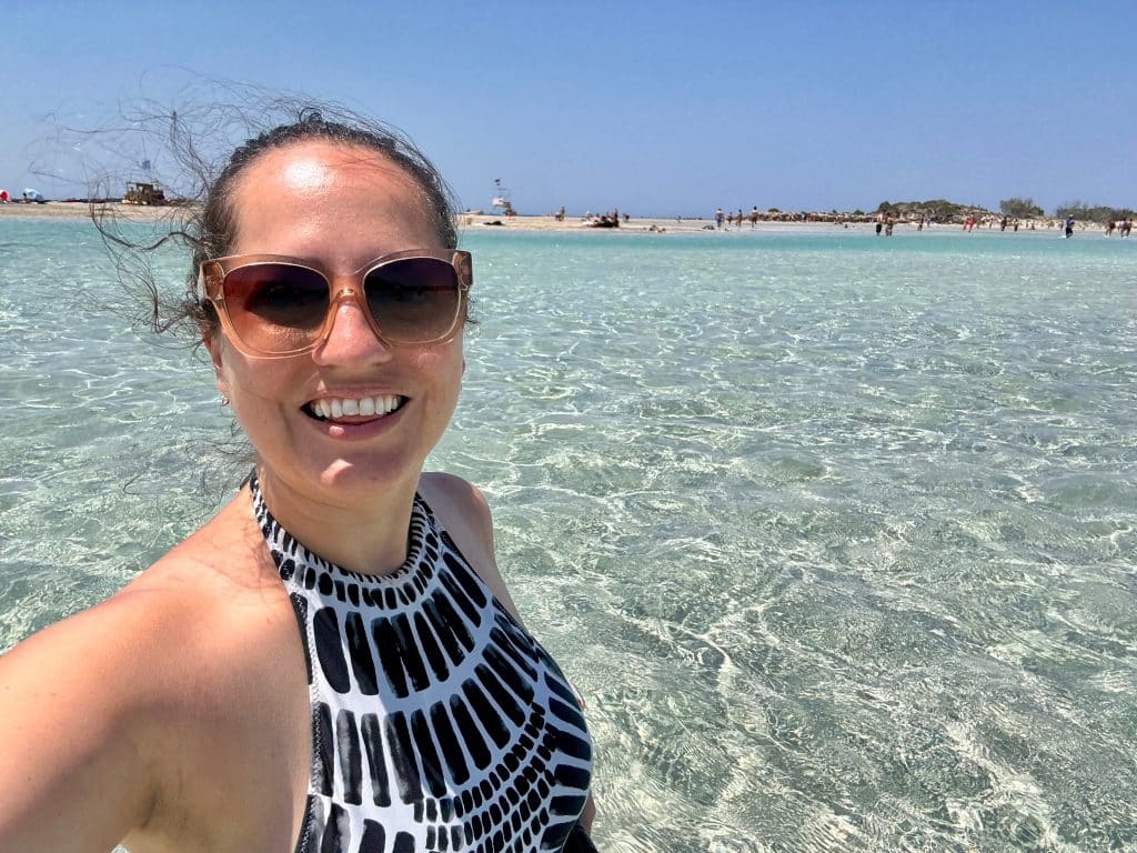 Kate taking a selfie in a black and white patterned bathing suit in front of a beach with incredibly clear turquoise water.